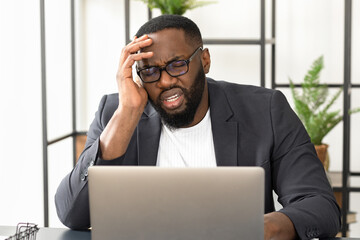 Tired African American businessman having headache or migraine sitting at the office desk. Young man freelancer sitting at home office suffers from excruciating pain in the temples