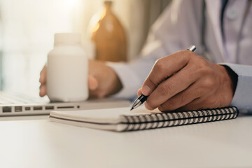 Pharmacist writing making notes in drug information using laptop computer sitting at desk. Woman physician, nurse or pharmacist wearing white coat writing in paper notebook. Close up view