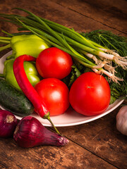 summer vegetarian still life - a set of vegetables for a vegan meal for the day. Fresh vegetables on a plate on a rustic wooden background, onions, tomatoes, cucumbers, peppers, garlic