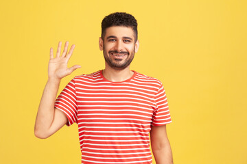 Hi you! Portrait of positive friendly bearded man in striped t-shirt saying hi raising hand, happy to see you, looking at camera with toothy smile. Indoor studio shot isolated on yellow background
