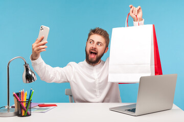 Positive bearded man blogger making selfie or recording video, posing at smartphone camera with shopping bags, bragging with purchases. Indoor studio shot isolated on blue background