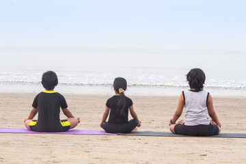 Back of boys and girl meditating in lotus position on mat practicing yoga at beach