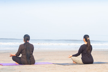 Fototapeta premium Rear view of two women meditating in yoga lotus position on exercise mat at beach