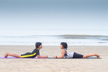 Profile of two boys practicing yoga cobra pose face to face on exercise mat at beach