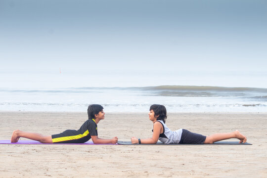 Profile of two boys practicing yoga cobra pose face to face on exercise mat at beach