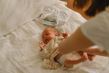 Happy young mother playing with baby while changing his diaper on bed. - obrazy, fototapety, plakaty