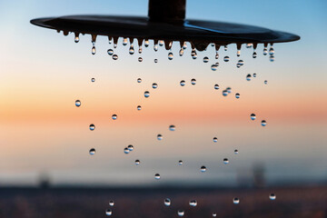 Water drops pouring from the outdoor beach shower head on blurred sunset seascape background,...
