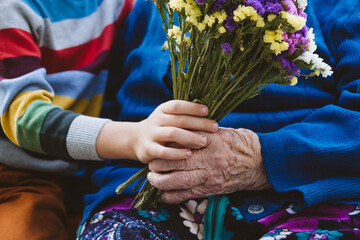 Close up female senior hand holding kid hand and flowers bouquet. Preschooler boy supporting his...