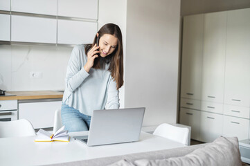 Smiling young woman using laptop computer for remote work from home, talks on the smartphone stands near desk and looks at the laptop screen, female freelancer has pleasant phone conversation