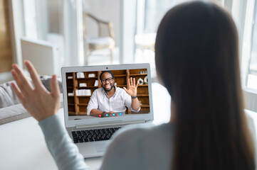 An African-American male employee, colleague or online tutor on the computer screen greeting, woman using laptop for video connection. Virtual conference, video call to coworker on the distance - obrazy, fototapety, plakaty