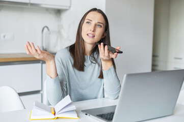 Smiling young woman is sending audio voice message by her smartphone working with laptop from home, female student using voice recognizable function for taking notes, searching information in network