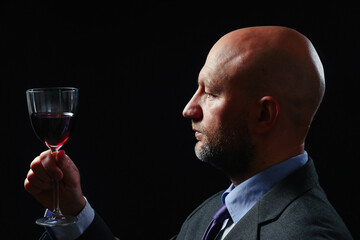 Portrait of a bald businessman with glass of red wine on dark background. Dramatic lighting. The model in in a suit in his 40s and grey beard.