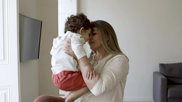 Cheerful mother enjoying sweet moments with son at home. Caucasian mom standing with little boy in living room, kissing and hugging him. Motherhood, tenderness, togetherness concept.
