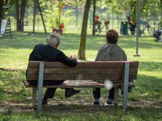 An Elderly Man and Woman Sitting on a Bench in a Public Park and Children's Games in the Background