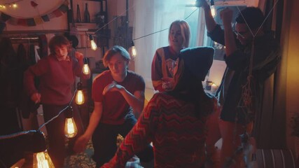 Handheld camera shot of young carefree men and women having drinks, smoking and dancing together on bed in retro room decorated with garland while having home party