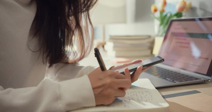 Close-up young asia lady freelance focus mobile phone chat with colleagues work finance writing on notebook in living room at home. Girl student learn online at house, Work from home concept.