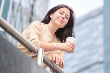 beautiful young woman enjoying a cup of coffee, against the backdrop of skyscrapers Smiling hipster girl with cup of hot beverage on city street.