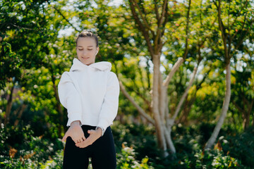 Horizontal shot of sporty European woman stretches hands and warms up before workout wears white sweatshirt andleggings poses against trees background spends free time outdoors on fresh air.