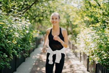 Sportive young woman has jogging workout training runs outdoor smiles pleasantly enjoys good day dressed in sportsclothes listens music from playlist. Start day from morning jog. Female runner