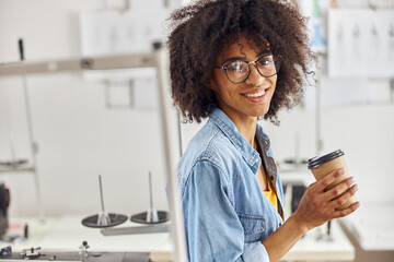 African-American seamstress with glasses and earring in nose holds cup near sewing machine in workshop