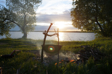 Camping in nature. Boiler hanging on a tripod over a campfire. Preparing dinner in a pot on the lake at sunset. Hike, travel concept.