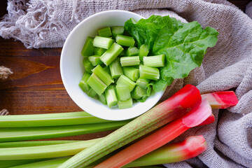 Red and green stems, stalks of rhubarb on a wooden surface. Rhubarb cut into pieces in a white bowl, green leaf. A fresh, eco-friendly plant.