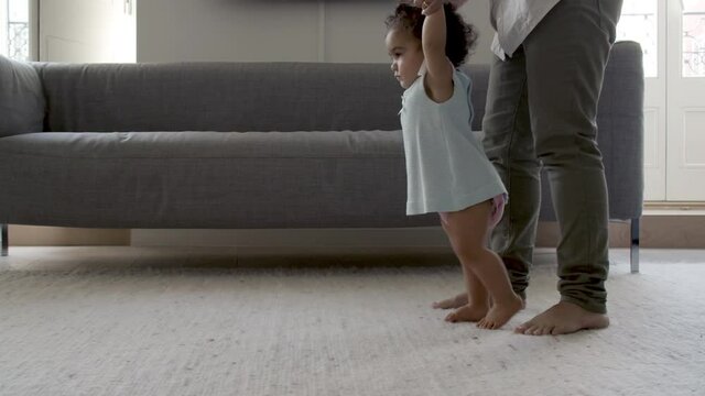 Focused little girl holding moms hands, doing efforts to walk. Cute curly-haired daughter taking her first steps at home, enjoying process of learning. First step, motherhood, nurture concept.