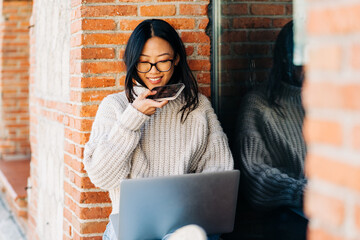 Smiling ethnic woman sending voice message on smartphone and working on laptop