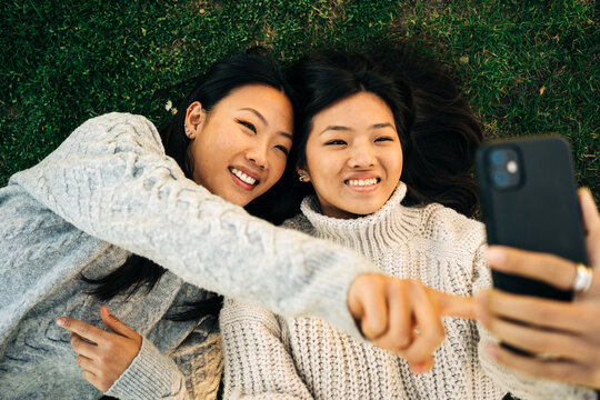 Joyful young ethnic female friends lying on lawn and showing tongues while taking selfie