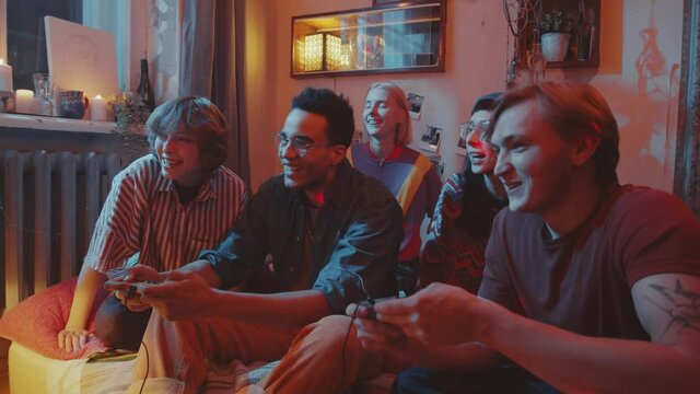 Company of young cheerful friends sitting together on bed in dark room with light projector, chatting and playing retro console game with excitement at home party