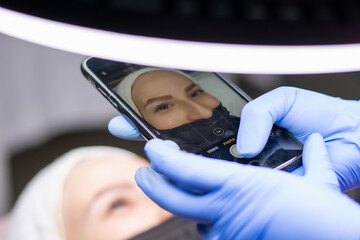 Unknown beautician's photographing the client girl's done eyebrows. Microblading and eyebrow tattoo concept. Beauty master in protective medical gloves holds phone. Close-up