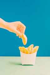 Modern healthy food summer idea with woman's hand holding a fresh organic orange slice. Fruit in french fries bag. Aesthetic vertical arrangement, creative colorful fast food concept.