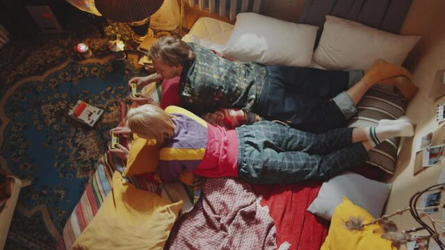 Top down shot of young man and woman lying on bed at home and using controllers while playing retro console game together