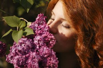 A beautiful young woman with red curly hair and freckels in a purple dress inhales the scent of the...