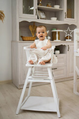 a cheerful little girl in a white dress is sitting in a high white chair in the kitchen. Food for children