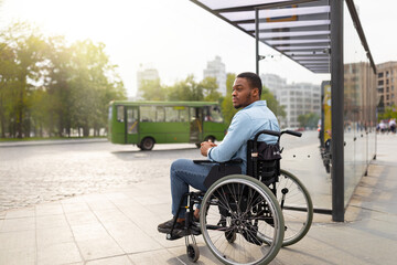 Young disabled black man in wheelchair waiting for public transport on bus stop, having difficulty...