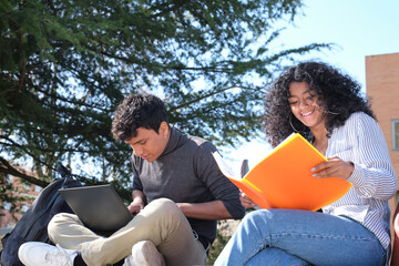 Two latin students studying from their lecture notes sitting on a wall outdoors. University life at...