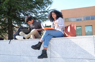 Two latin students studying from their lecture notes sitting on a wall outdoors. University life at campus.