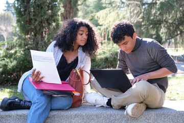 Two latin students studying together sitting on a bench outdoors. University life at campus.