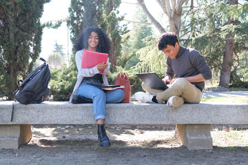 Two latin students smiling studying together sitting on a bench outdoors. University life at campus.