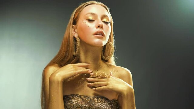 Portrait of beauty woman, face, body in gold paint. Golden shiny skin. Fashion model blonde girl posing in studio. Glamorous earrings, ring jewellery accessories. Professional metallic glitter makeup.