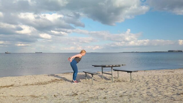 Young caucasian woman doing gym exercises and training on the beach in Amager, Copenhagen, Denmark. The girl is doing box jumps on a bench. It is a sunny spring evening. 3x times speeded up 4K clip