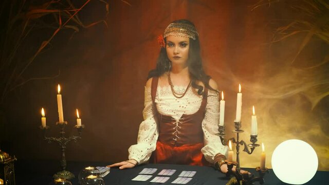 Fantasy beautiful girl in image of gypsy witch in dark gothic room. Art dress red costume. Fortune teller magical woman reading future on tarot cards crystal ball. Ritual candles burning, seance smoke