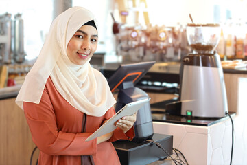Religious asian muslim woman in orange dress and white shaft standing and holding tablet with confidence. Business woman stand and looking camera with coffee shop counter blurred background
