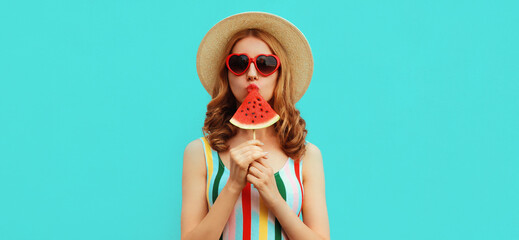 Summer portrait of young woman sucking ice cream shaped slice of watermelon wearing a straw hat, red heart shaped sunglasses on blue background