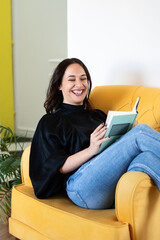 Smart Young Woman Gains Knowledge By Reading A Book. Brunette Spends Her Leisure Reading Book While Sitting In Cozy yellow Armchair.