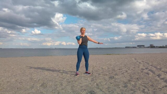 Young caucasian woman doing gym exercises and training on the beach in Amager, Copenhagen, Denmark. The girl is training with a kettle ball. It is a sunny spring evening. Real time 4K