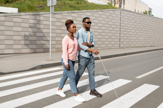 Young Black Woman Assisting Visually Impaired Millennial Guy With Cane Crossing City Street. Vision Disability Concept