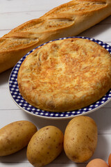 Tortilla de Patatas española. Delicioso plato salado basado en Huevo, Patatas y a veces Cebolla. Acompañada por patatas, una barra de pan, y huevos. En fondo de madera blanca, entera y sin cortar. 