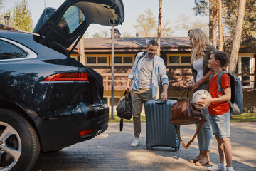 Happy young family with little packing stuff into the car and smiling while standing near house